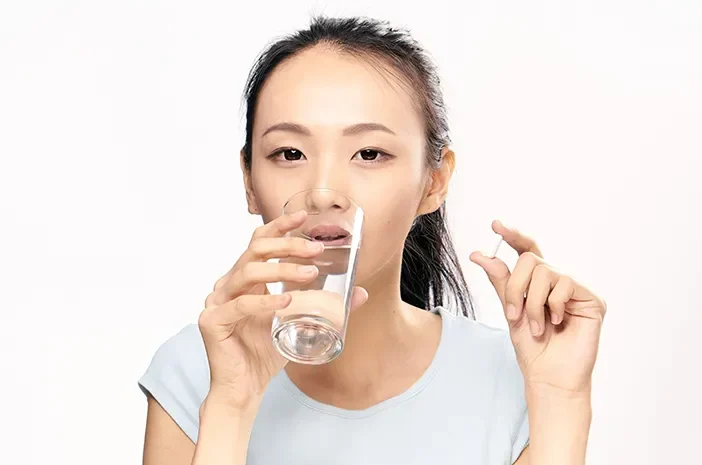 Wanita memegang pil dan gelas air putih, mengindikasikan konsumsi suplemen.