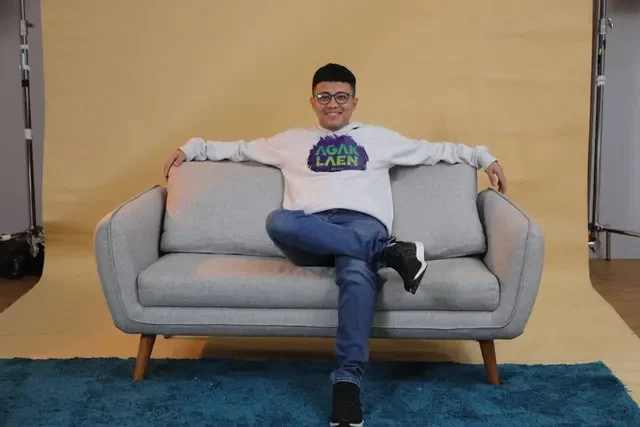 Bene Dion, salah satu pemeran utama film Agak Laen, duduk santai di sofa.