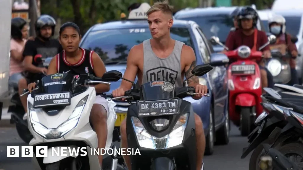 Pria dan wanita mengendarai skuter di jalan raya padat, dikelilingi kendaraan lain.