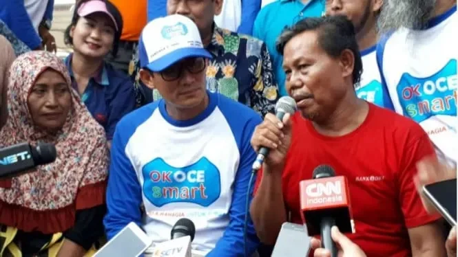Kerumunan orang dengan mikrofon di acara publik dengan beberapa orang yang berbicara.
