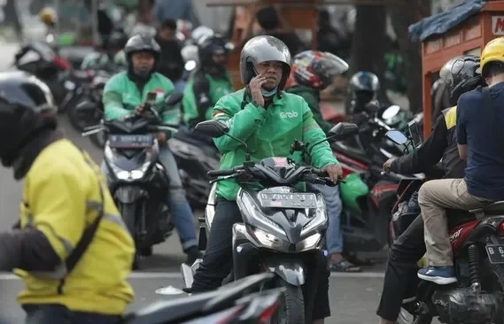 Pengemudi ojek online menggunakan ponsel sambil mengendarai motor di tengah keramaian.