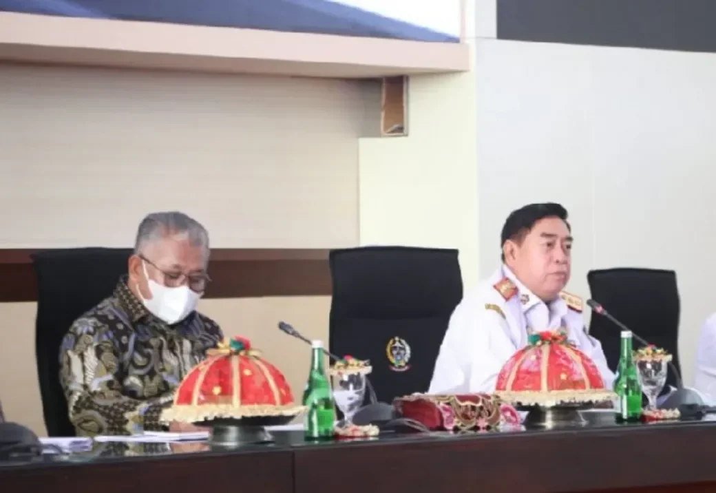 Dua pejabat pria dalam pakaian formal, satu bermasker, saat menghadiri pertemuan.