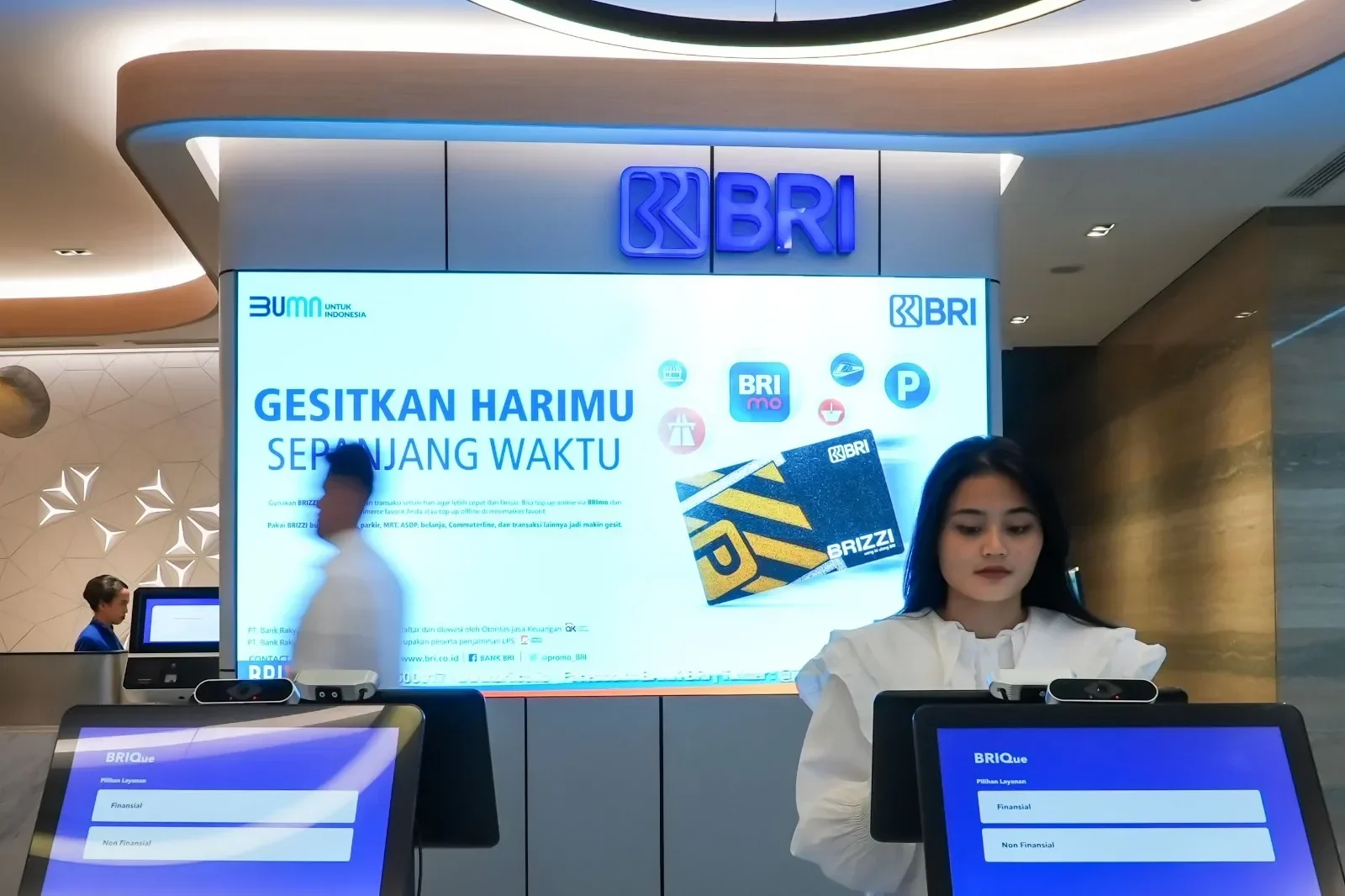 Petugas bank BRI melayani nasabah di depan layar digital promo BRIZZI dan BRImo.