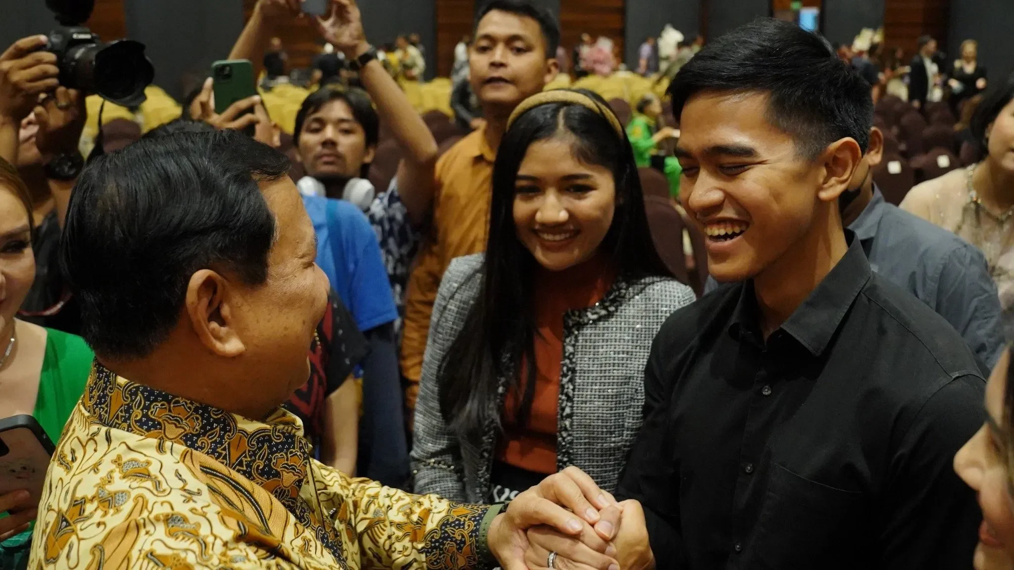 Prabowo Subianto berjabat tangan dengan seorang pria di tengah kerumunan, disaksikan wanita dan fotografer.