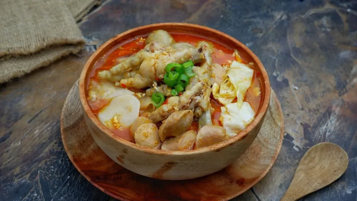 Semangkuk seblak berisi kerupuk, ceker ayam, bakso, sosis, dan sayuran, berkuah pedas, dalam mangkuk kayu.