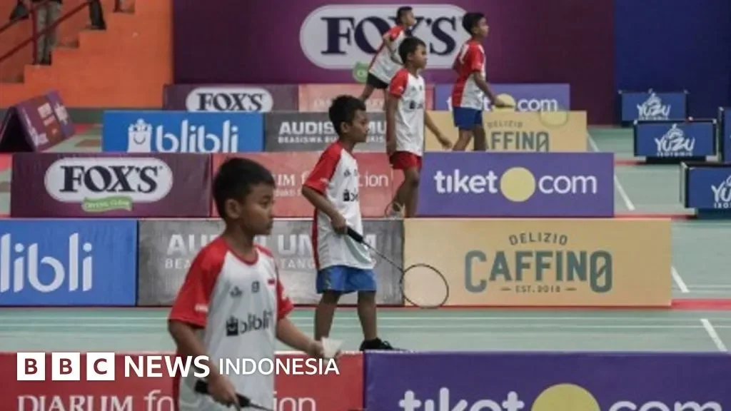 Anak-anak bulu tangkis berlatih di turnamen junior dengan sponsor di latar belakang.