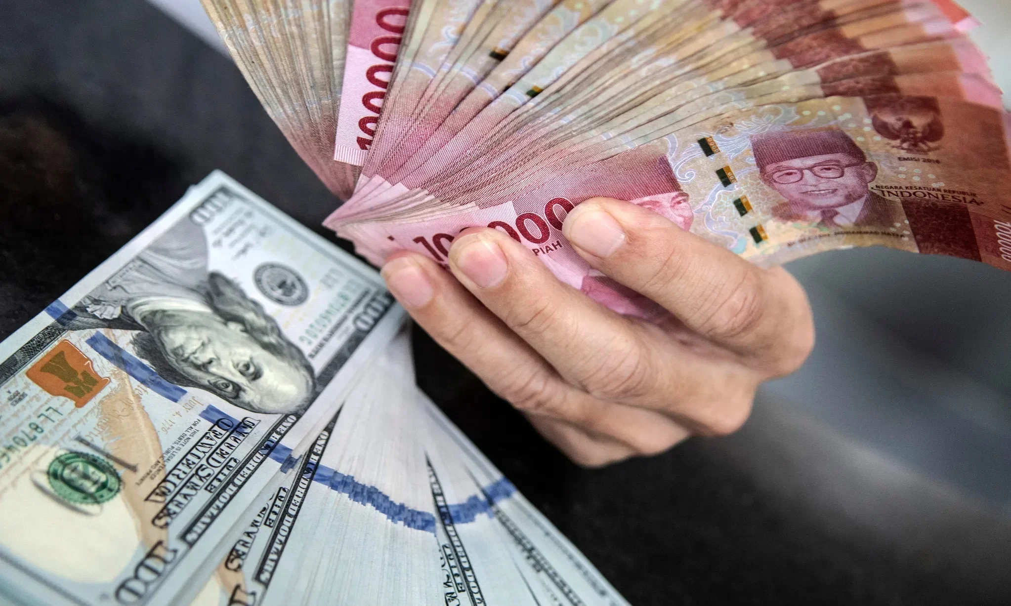 Tangan memegang uang Rupiah di atas tumpukan uang Dolar AS.
