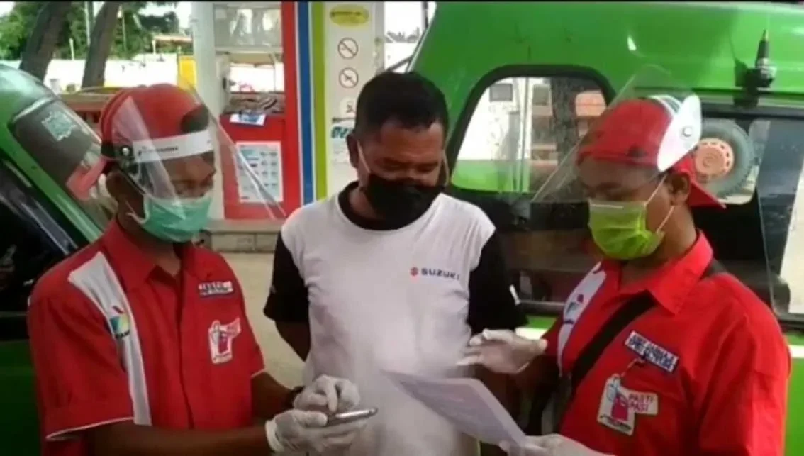Petugas SPBU berbaju merah dan pria berkaus Suzuki berdiskusi di dekat mesin pompa BBM.