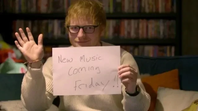 Ed Sheeran memegang papan bertuliskan "New Music Coming Friday" sambil melambai.