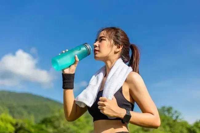 Wanita minum air dari botol setelah olahraga, langit biru cerah.