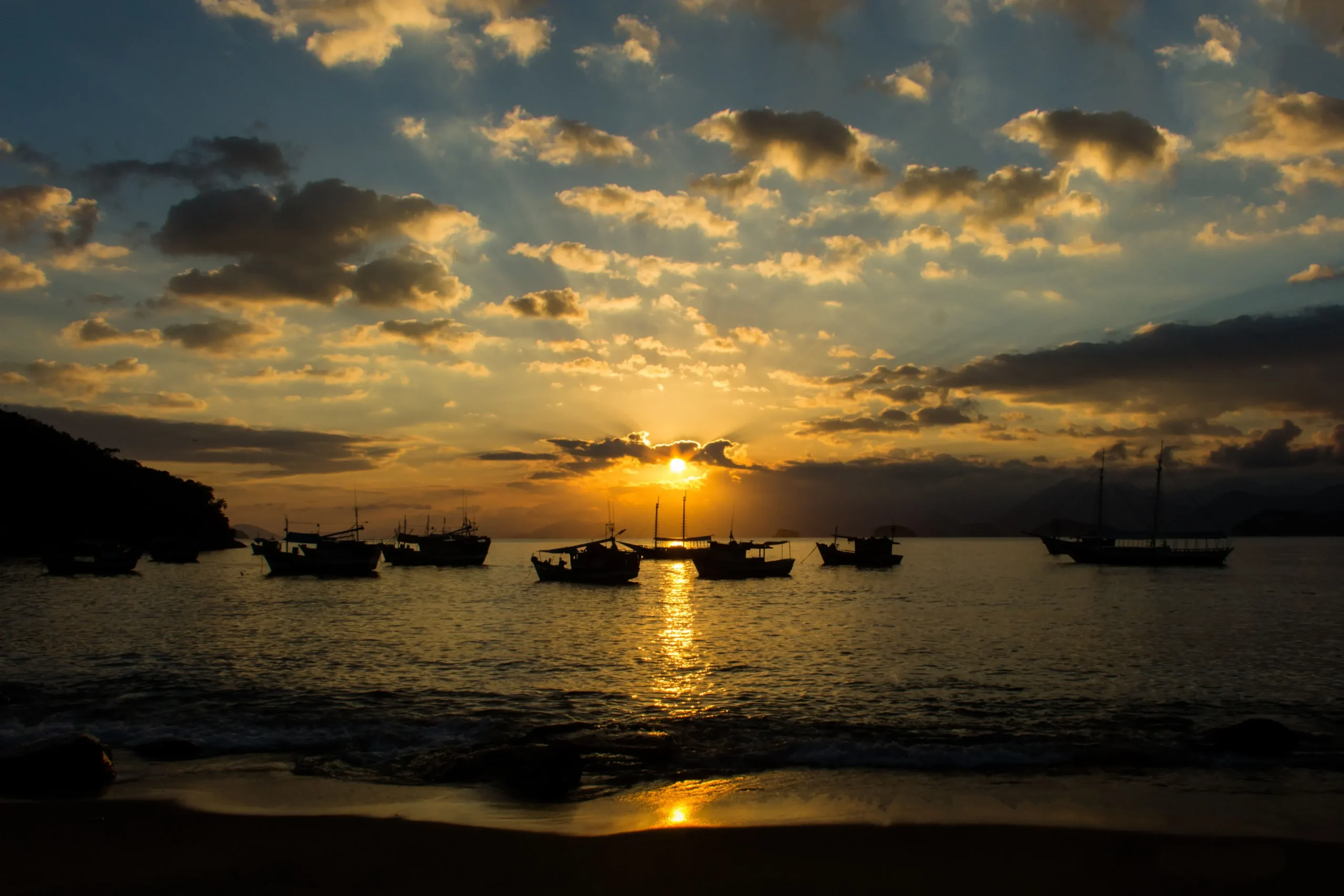 Siluet perahu di laut saat matahari terbenam dengan pantulan cahaya keemasan di permukaan air.