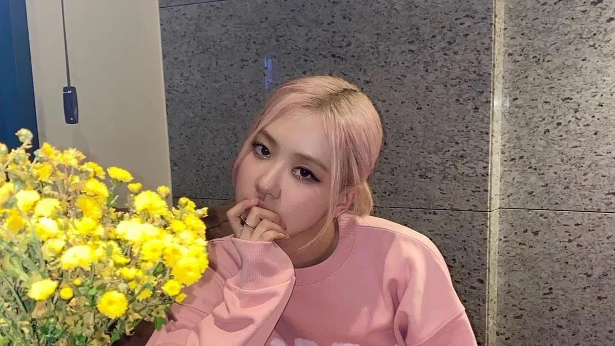 Wanita muda berambut pink memakai sweter merah muda, di samping buket bunga kuning.