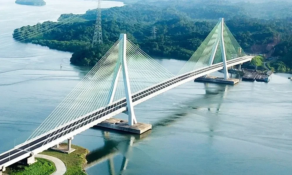 Jembatan kabel-tetap modern melintasi perairan, dikelilingi perbukitan hijau, simbol kemajuan infrastruktur.