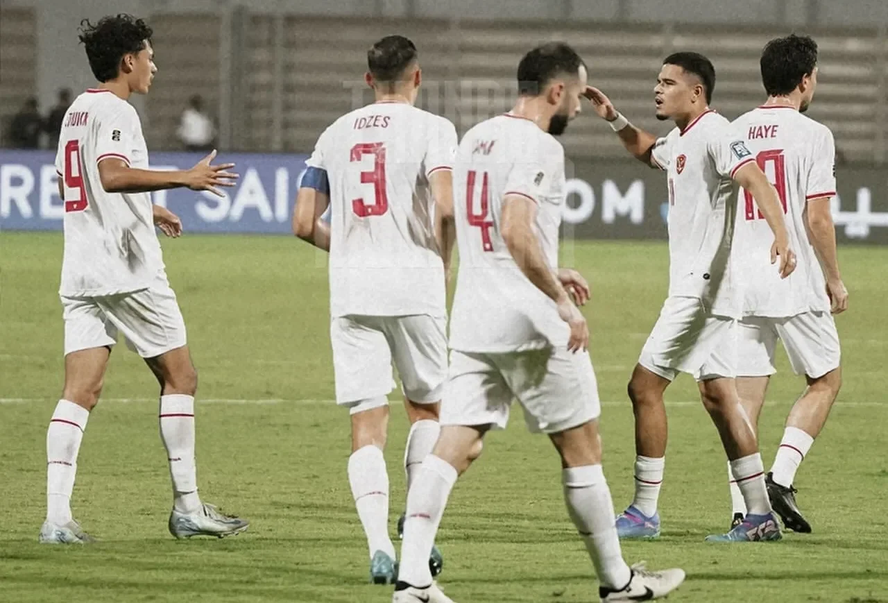 Beberapa pemain sepak bola berjersey putih sedang berinteraksi di lapangan hijau.
