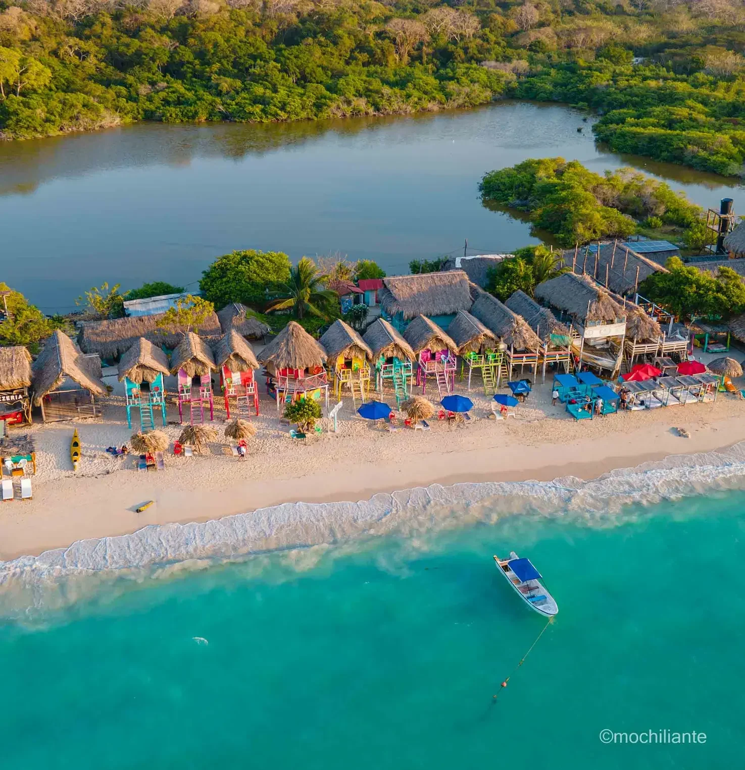 Pemandangan pantai tropis dengan pondok warna-warni dan perahu di laut biru kehijauan.