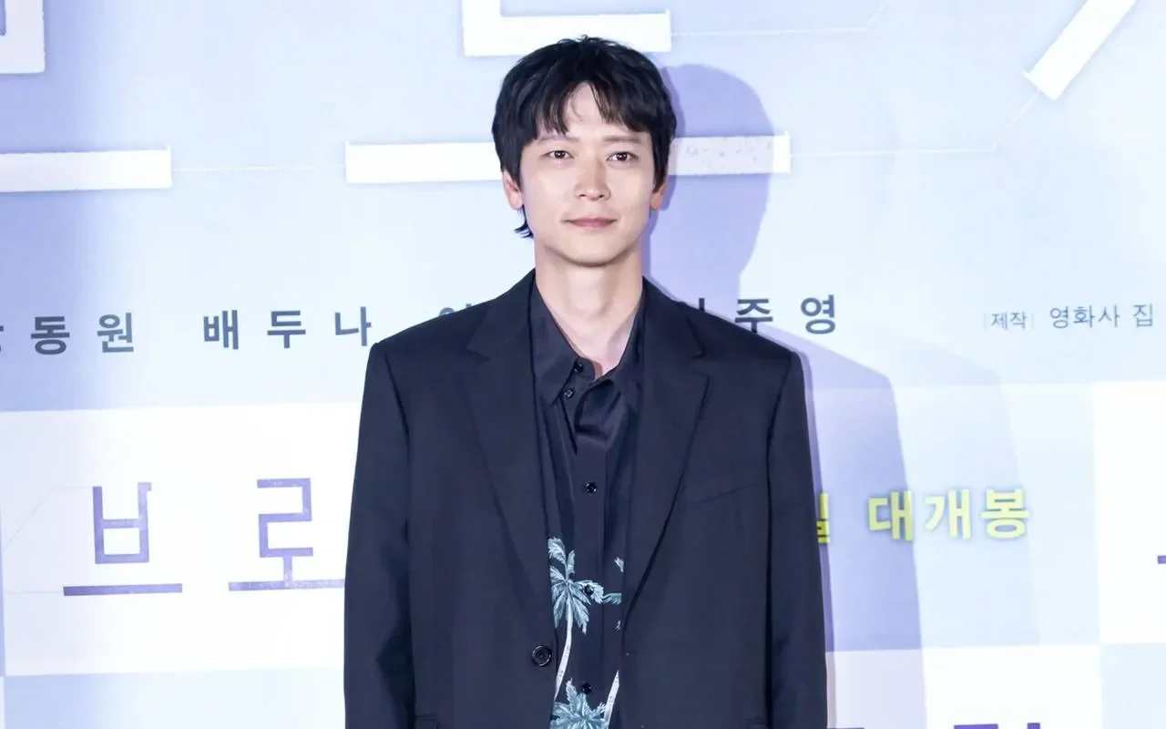 Aktor Gang Dong-won mengenakan jas hitam dengan kemeja bermotif pohon palem di depan latar belakang biru muda.