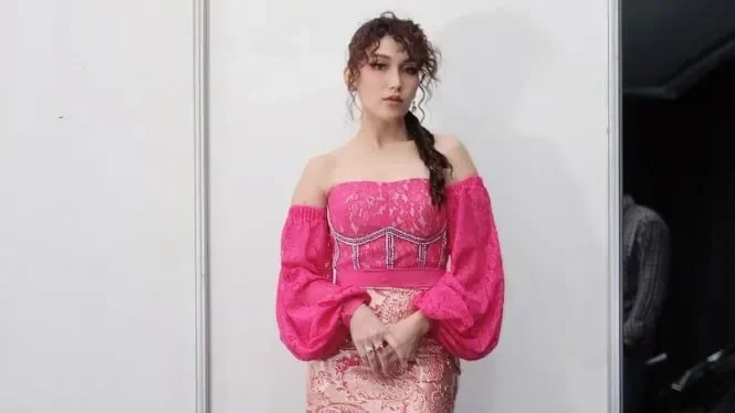 Seorang wanita bergaun merah muda, rambut dikepang, berpose dengan ekspresi serius.