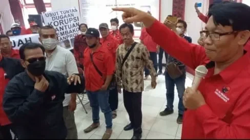 Suasana tegang sebuah pertemuan partai. Seorang pria berbicara dengan mikrofon, disaksikan kerumunan peserta.