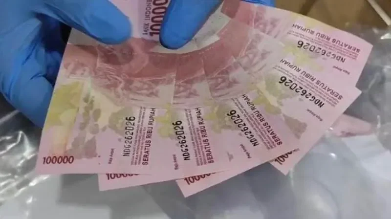 Uang pecahan seratus ribu rupiah dipegang oleh tangan bersarung tangan biru.