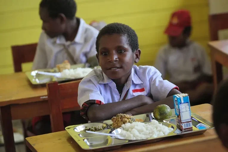 Seorang siswa sekolah dasar dengan nampan makanan di depannya, tampak menanti.