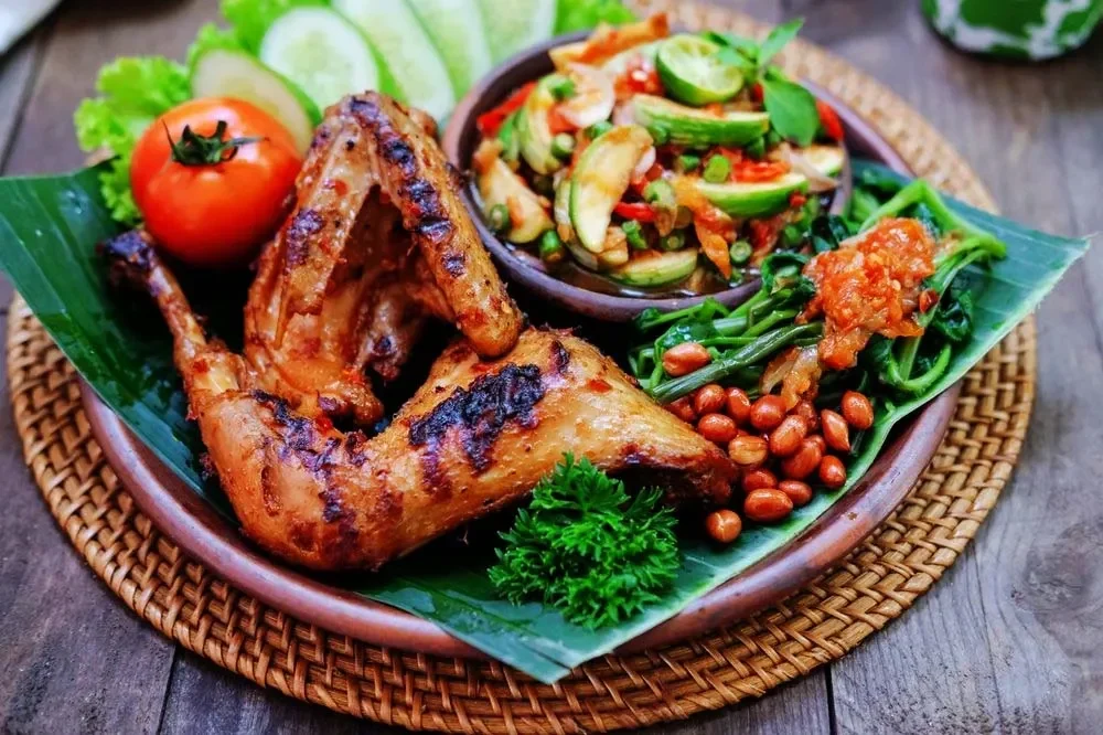 Sajian ayam bakar utuh dengan kulit kecoklatan, dilengkapi sambal pedas dan lalapan segar.
