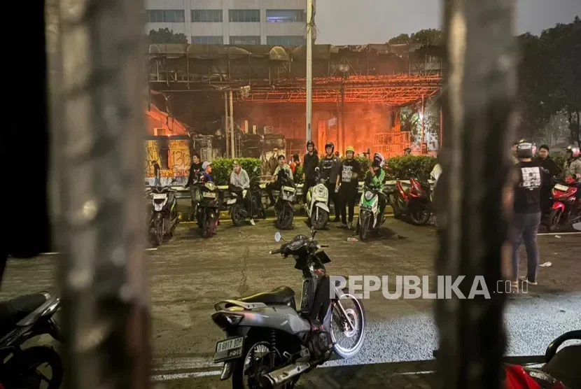 Sekelompok pemuda berkumpul dengan motor di depan area yang terbakar dan puing-puing.