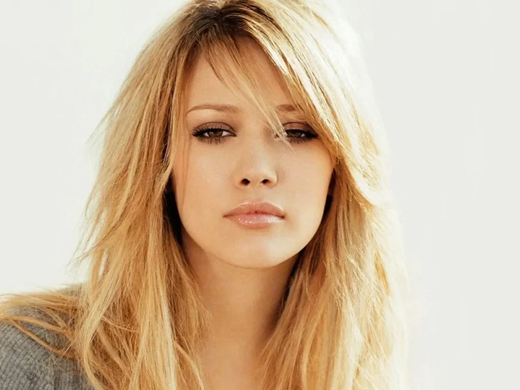Foto close-up Hilary Duff dengan rambut pirang bergelombang dan riasan glamor.