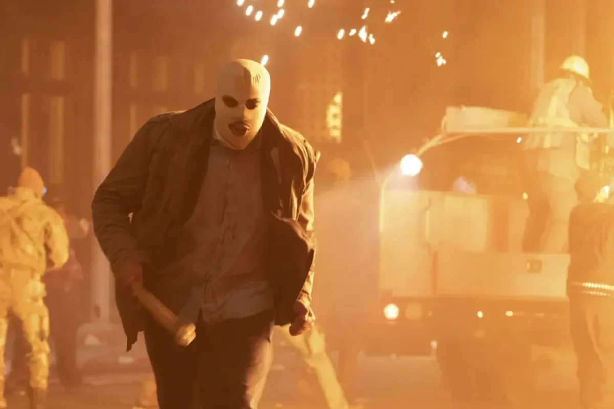 Pria bertopeng menyerang di tengah kerusuhan dengan latar belakang api dan kendaraan.