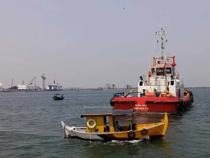 Kapal nelayan kecil dan kapal tunda beroperasi di perairan pelabuhan yang sibuk.
