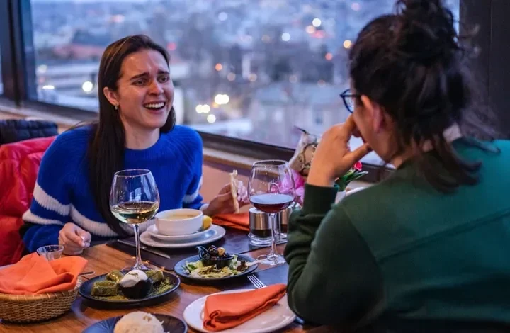 Dua wanita makan malam dan tertawa di restoran dengan pemandangan kota.