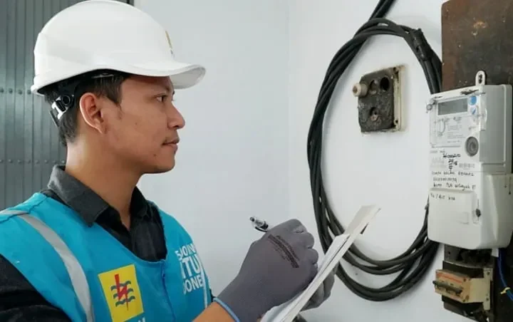 Teknisi PLN memeriksa meteran listrik sambil memegang dokumen.