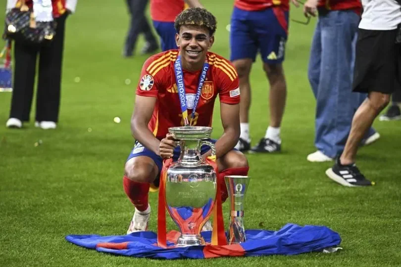 Pemain muda Spanyol memegang trofi, latar belakang kerumunan dan rumput hijau.