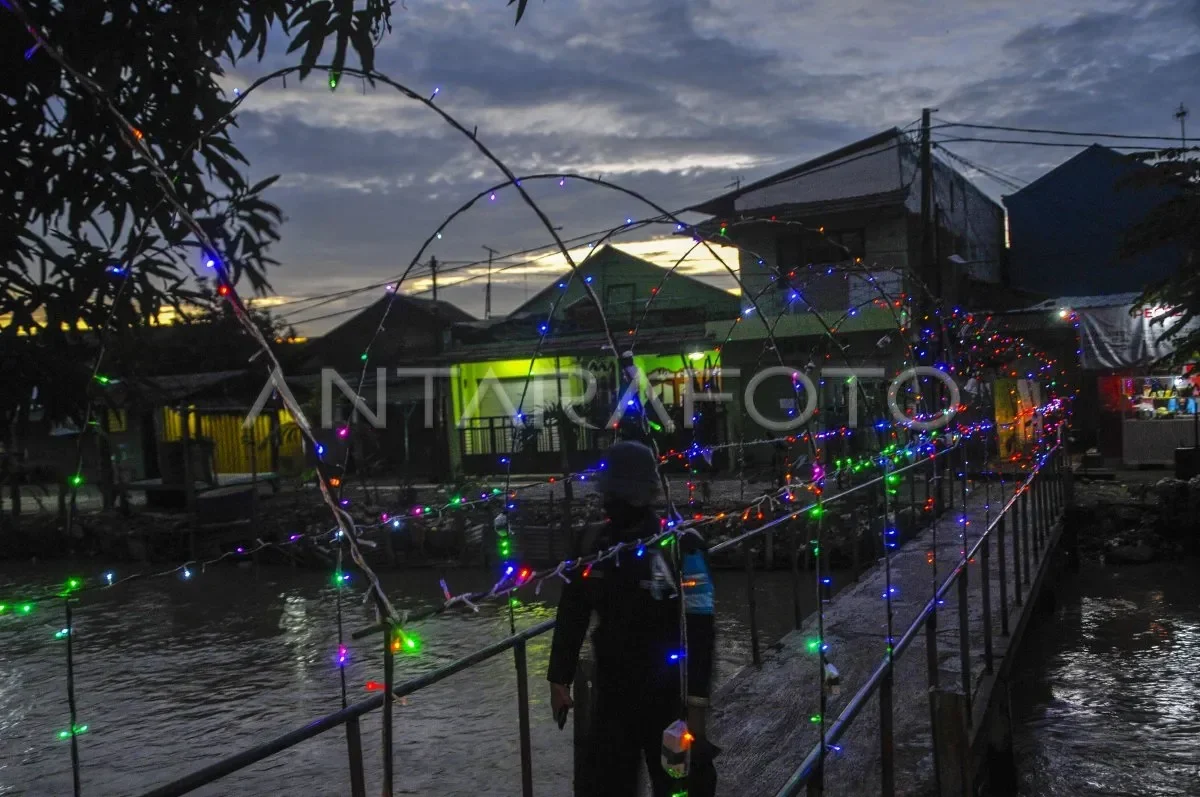 Seorang warga melintasi jembatan di malam hari, dihiasi lampu warna-warni di tengah pemukiman.