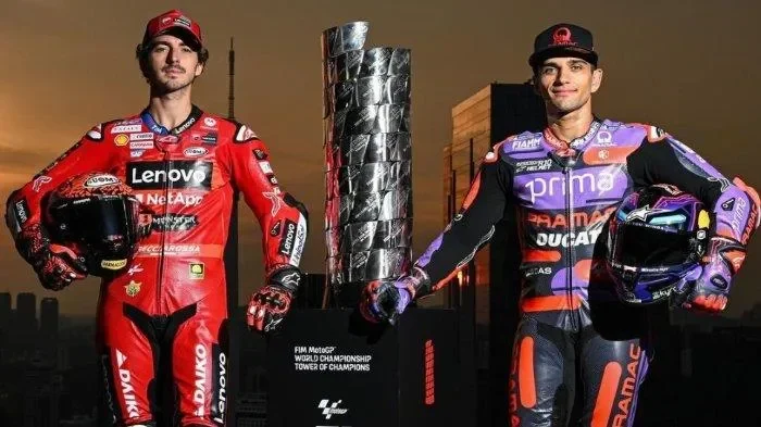 Dua pembalap MotoGP berdiri di dekat trofi juara dunia MotoGP dengan latar belakang perkotaan.