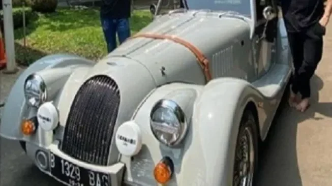 Mobil klasik berwarna putih dengan plat nomor Jakarta, detail lampu dan gril.