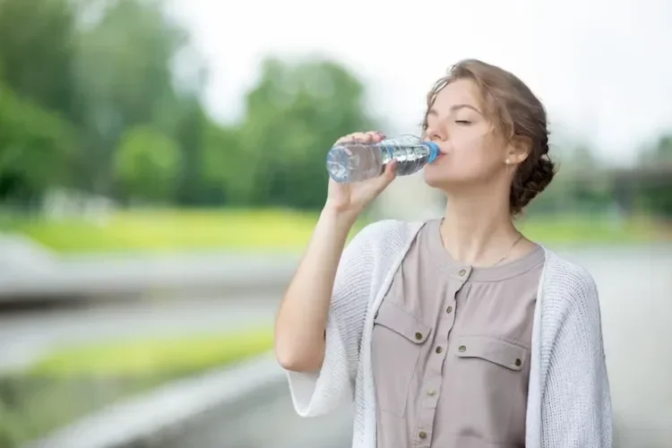 Seorang wanita minum air dari botol plastik sambil berdiri di luar.