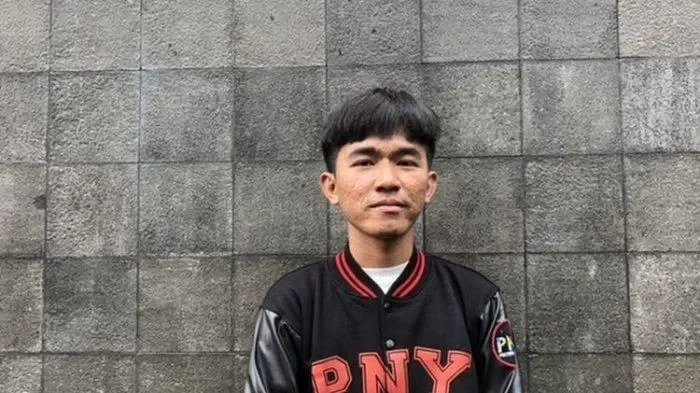 Pria berjaket hitam berpose di depan dinding batu, kemungkinan saksi.