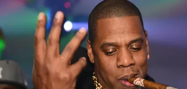 Penyanyi Jay-Z memegang cerutu, tangan terangkat di tengah keramaian.