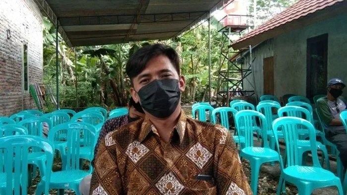 Potret suasana duka, seorang pria memakai batik dan masker di tengah area berkumpul.