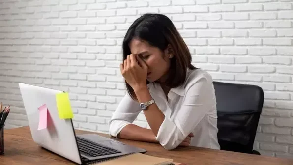 Wanita lelah memegang kening di meja kerja, menggambarkan efek jetlag yang menyiksa pikiran.