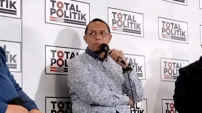 Hasan Nasbi berbicara memegang mikrofon dengan latar belakang logo Total Politik.