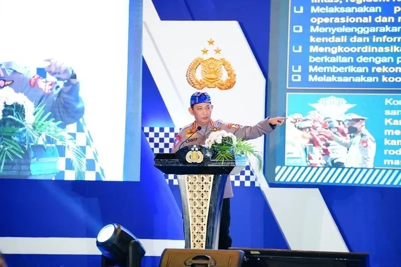 Pejabat Polisi memberikan pidato di podium, menunjuk ke layar dengan logo polisi.