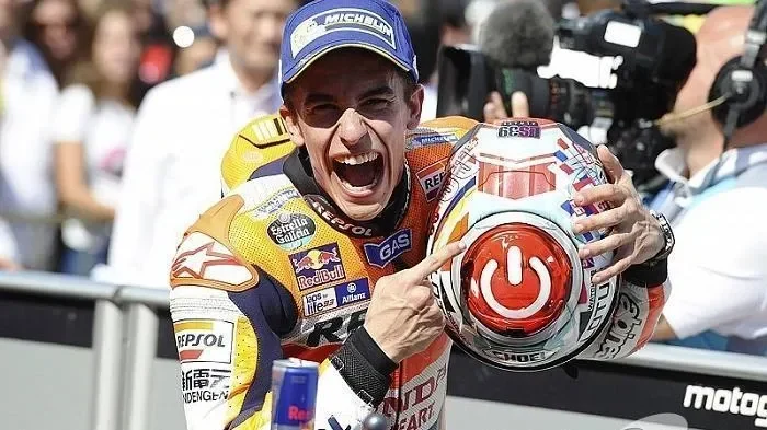 Marc Marquez merayakan kemenangan dengan ekspresi gembira, mengenakan topi dan baju balap, menunjuk helmnya.