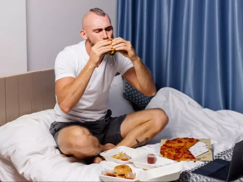 Pria makan burger di ranjang dengan pizza dan junk food lainnya.