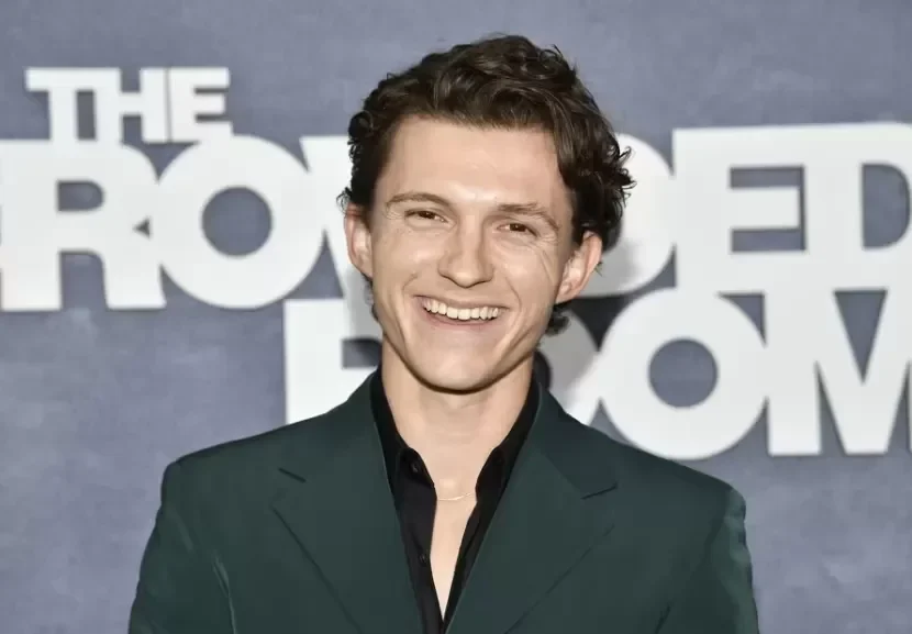Tom Holland tertawa mengenakan jas hijau tua.