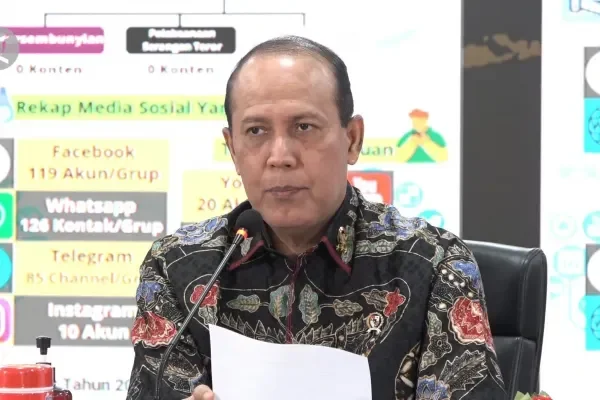 Pria berbatik berbicara di mikrofon dengan tampilan data rekap media sosial di latar belakang.