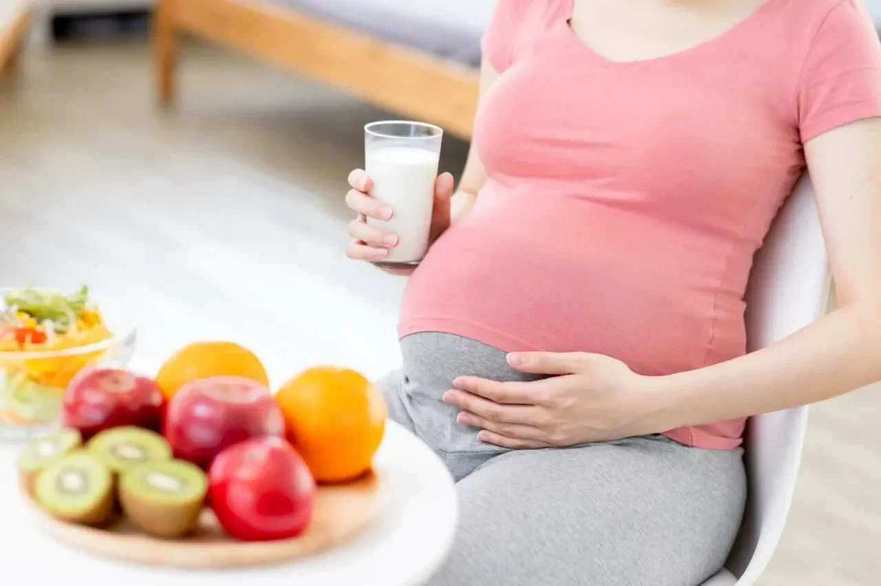 Ibu hamil memegang segelas susu, dengan buah-buahan dan salad segar di meja.