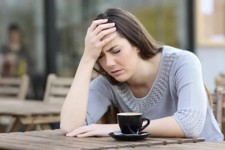 Wanita tampak pusing dengan secangkir kopi di meja kayu luar ruangan.