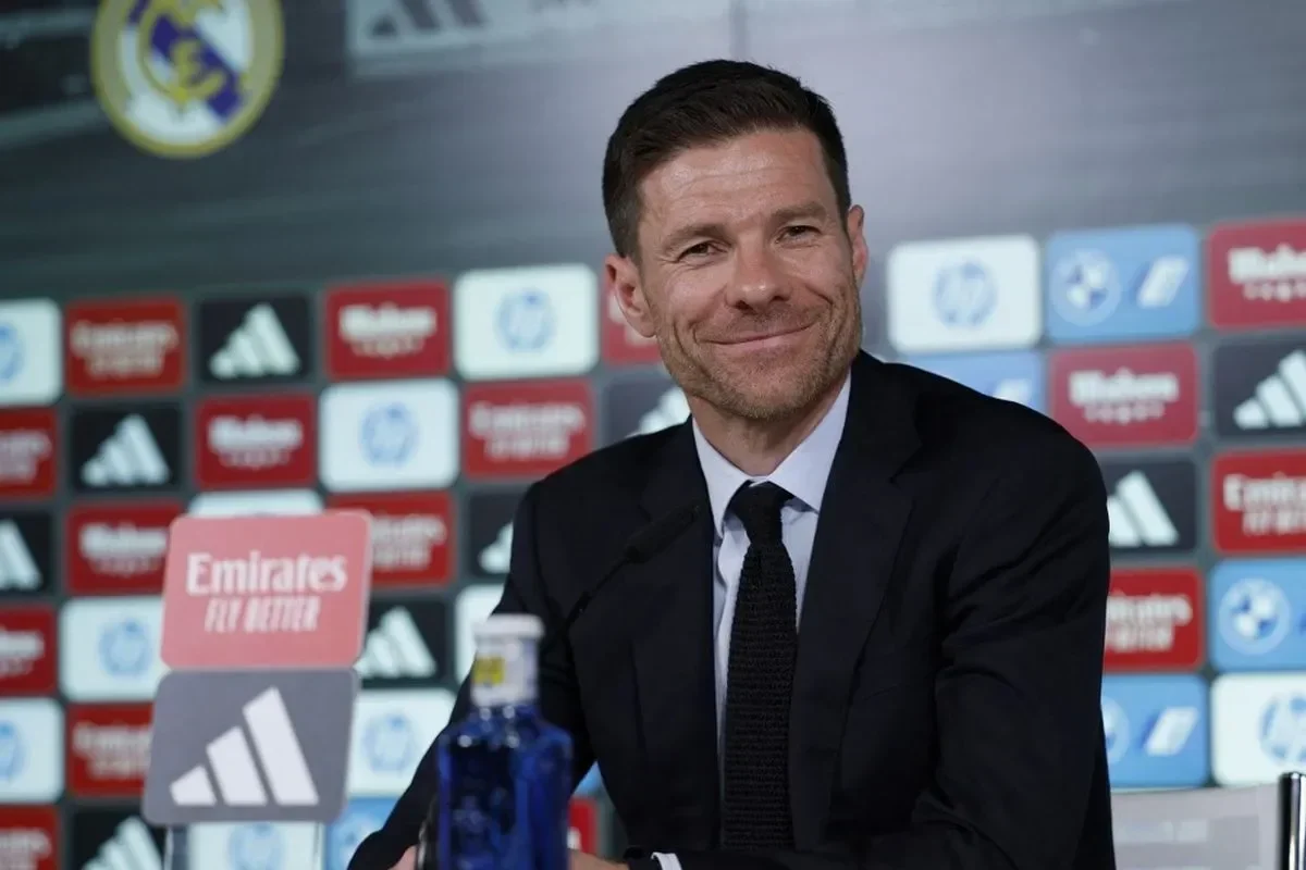 Xabi Alonso tersenyum bahagia saat konferensi pers, mengenakan jas hitam dengan dasi rajut.