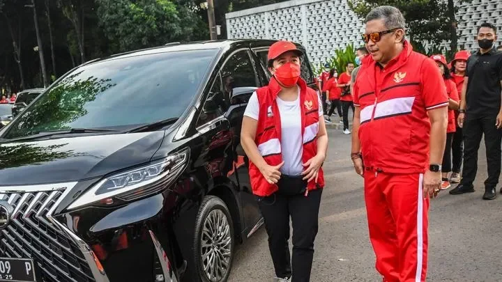Dua politisi berseragam merah di depan mobil mewah, kerumunan di belakang.